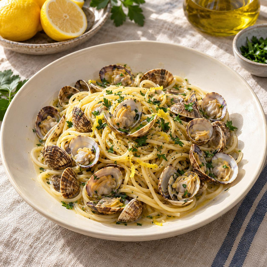 Dorset clams with spaghetti, parsley and lemon zest in a wide cream bowl on linen with halved lemons behind