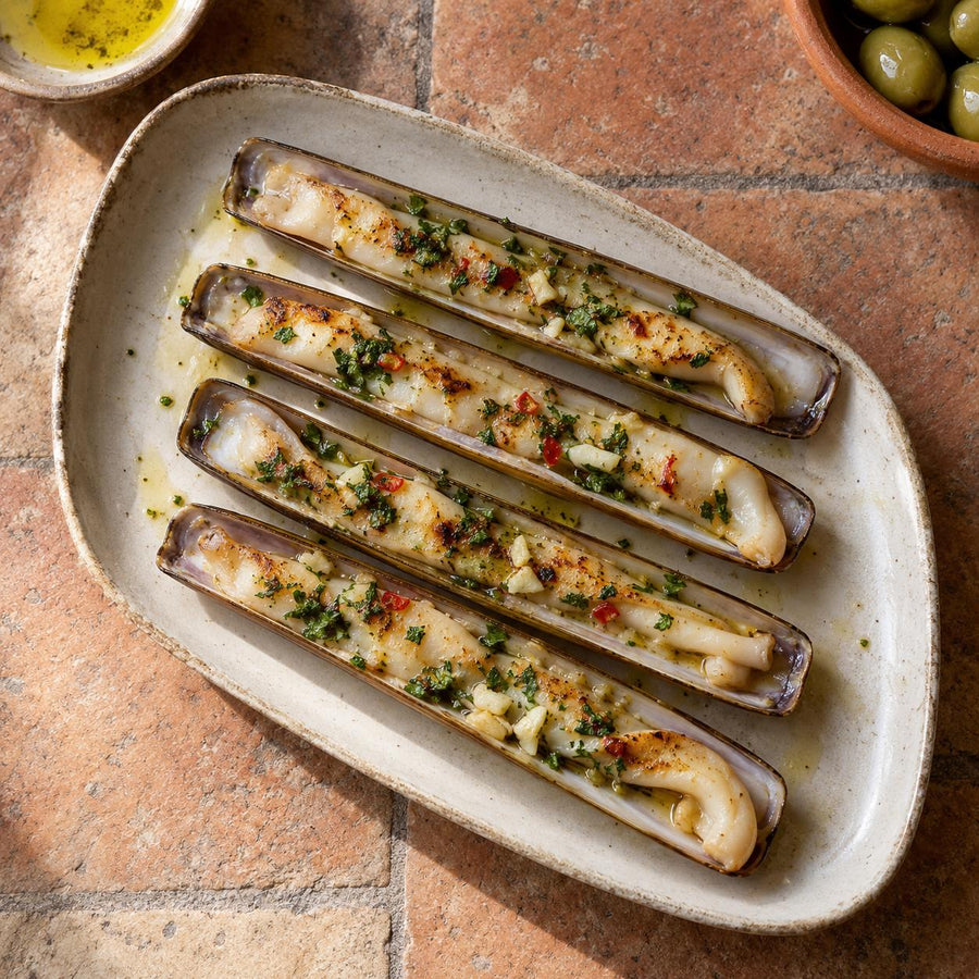 Grilled razor clams on the half shell with garlic, parsley and red chilli in olive oil on a cream stoneware platter