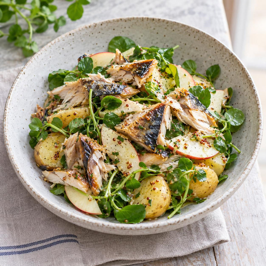 Smoked mackerel fillets flaked over new potatoes, apple slices and watercress with mustard dressing in a stoneware bowl