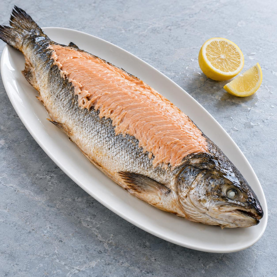 A whole dressed salmon on a white oval platter with lemon on a grey stone surface